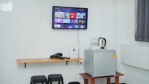 Una habitación pequeña y moderna con un televisor de pantalla plana montado en la pared que muestra diversas aplicaciones de transmisión. Hay un refrigerador pequeño sobre un soporte de madera, una estantería con un teléfono y dos taburetes negros.