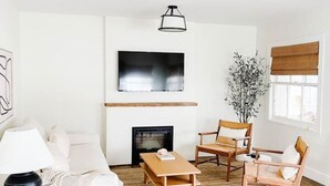 Ein Wohnzimmer mit einer Couch, zwei Sesseln, einem Couchtisch, einem Kamin und einem an der Wand befestigten Fernseher.