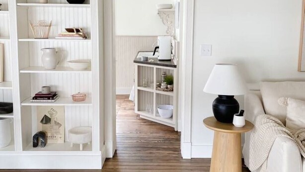 Ein Zimmer mit einer weißen Regalkonstruktion, die mit verschiedenen Gegenständen gefüllt ist, einem Holzboden, einem runden Beistelltisch mit Lampe und einem beigen Sofa.