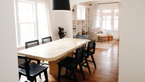 A dining area with a large table and chairs, a black hanging light fixture, and a bookshelf in the background.