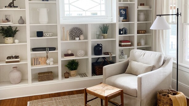 A living room with a white sofa, a small wooden table, and a floor-to-ceiling white shelving unit filled with various decorative items.