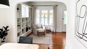 A spacious room with a large white bookshelf, a comfortable armchair, a small table, and a view of the outdoors through a window.