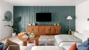 En moderne stue med en grøn accentvæg, et fladskærms-tv, et træskab og et hyggeligt opholdsområde med en sofa og et sofabord.