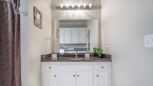 Une salle de bain avec des meubles blancs, un comptoir en granit, un miroir et un rideau de douche.