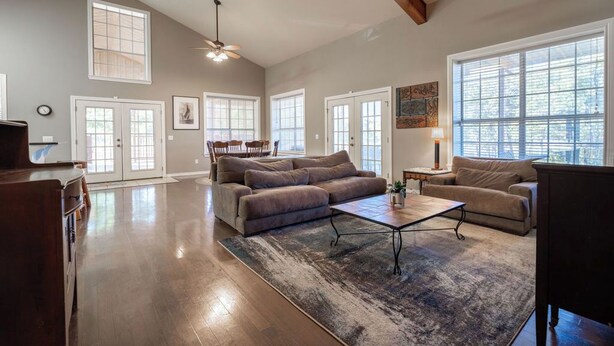 Un salon spacieux avec un grand canapé, une table basse et un ventilateur de plafond. De nombreuses fenêtres laissent entrer la lumière naturelle et offrent une vue sur l’extérieur.