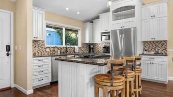 A kitchen with white cabinetry, a central island with bar stools, a stainless steel refrigerator, and a window with a view of greenery.