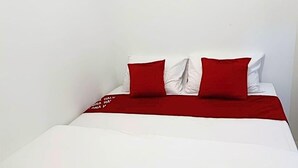 Un lit avec une literie blanche et des oreillers rouges, un mur blanc et un tableau encadré au-dessus du lit.