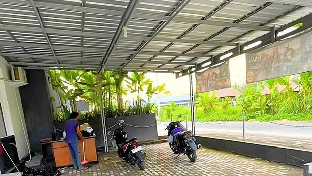 Un espace de stationnement couvert où sont garées deux motos, un bureau avec une chaise, et un sol carrelé présentant des fissures apparentes.
