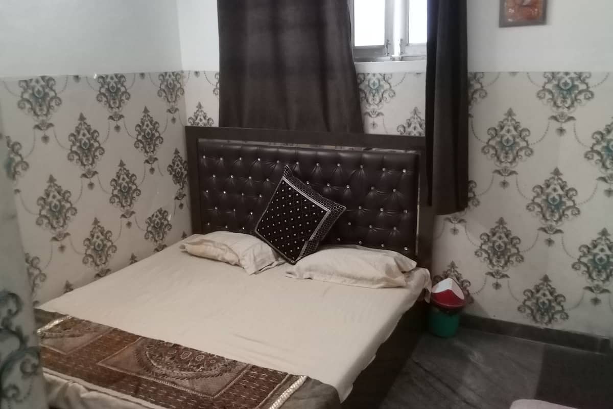 A bedroom with a bed, a window with curtains, patterned wallpaper, and a small trash bin.
