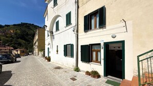 A narrow cobblestone street with a building featuring a white facade, green shutters, and a visible door with a sign.