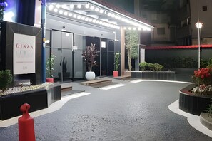 Entrada de hotel con diseño moderno, fachada de vidrio, plantas en macetas y un hidrante rojo.