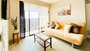 A living room with a sofa, a coffee table, a TV mounted on the wall, a potted plant, and a cityscape view through the window. - R Lindo departamento 1BR en Miraflores (Lima)