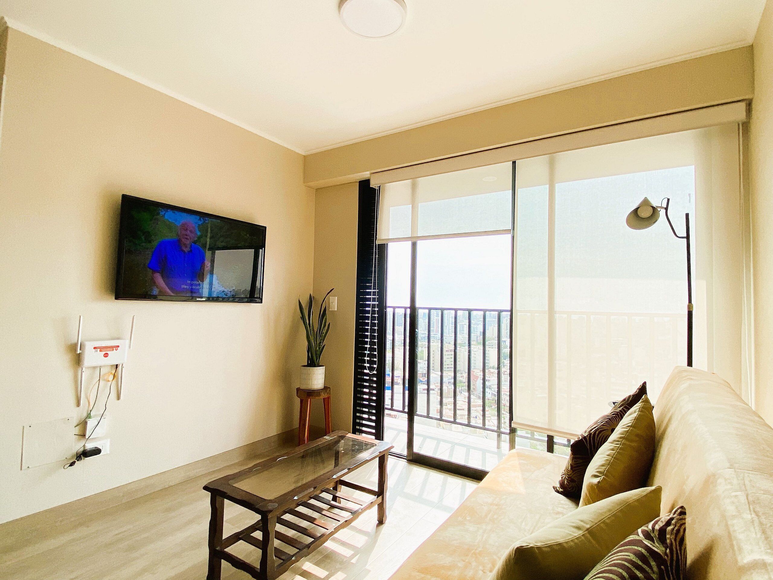 A living room with a sofa, a glass-top coffee table, a TV mounted on the wall, and a balcony with a view of the city.