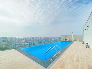 A rooftop pool with a city view, a wooden deck, and a glass railing.
