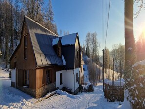 Una casa de madera con techo inclinado, rodeada de terreno y árboles cubiertos de nieve.