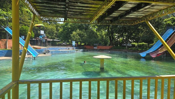 Una piscina con scivoli, circondata da alberi e una zona coperta con tetto.