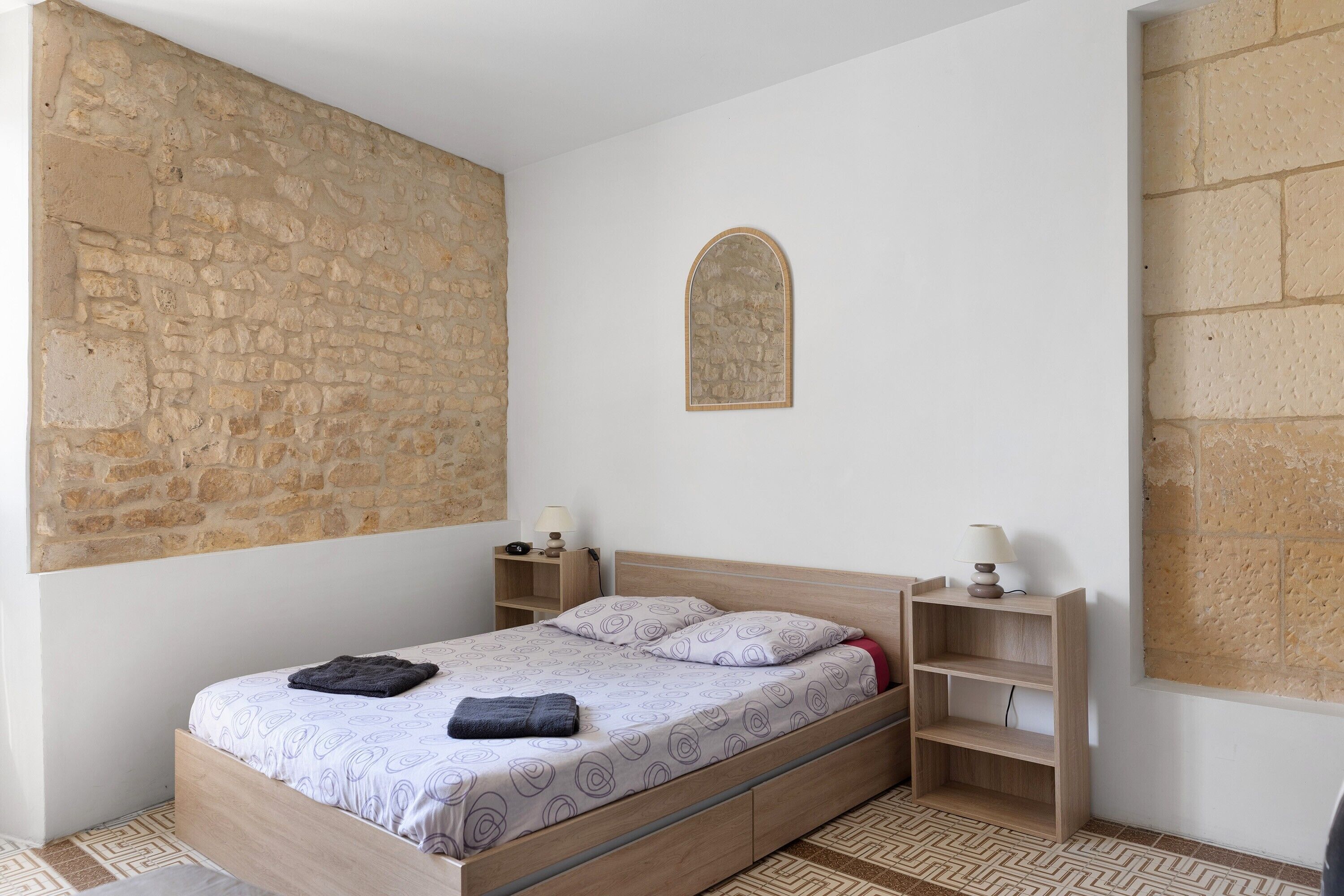 Une chambre avec un mur en pierre, un lit en bois, une petite table de chevet, une lampe et une étagère.