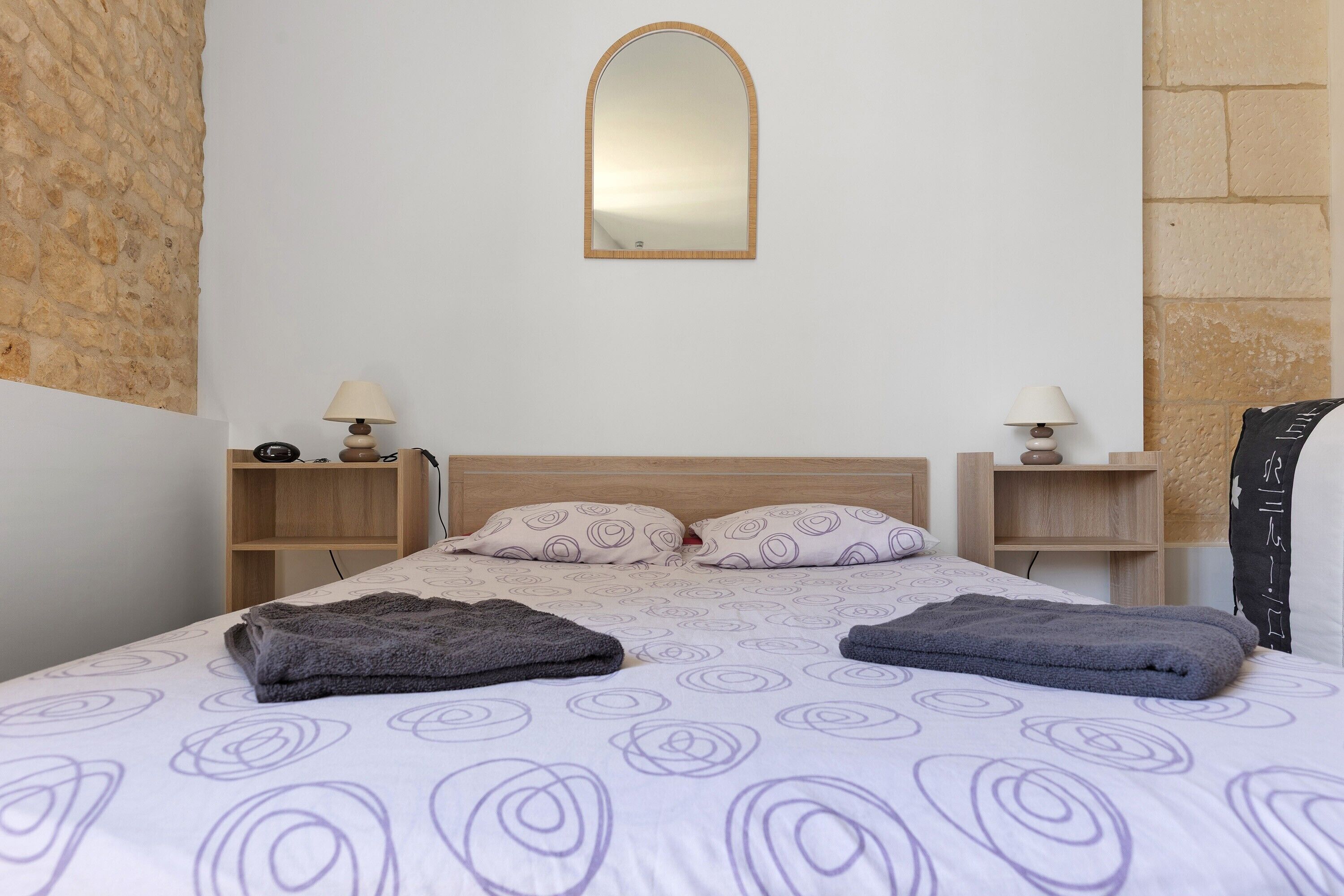 A bedroom with a bed, two bedside tables, a mirror, and a stone wall.