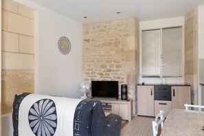 A room with a stone wall, a flat-screen TV, a fan, a table with chairs, and a window with blinds.