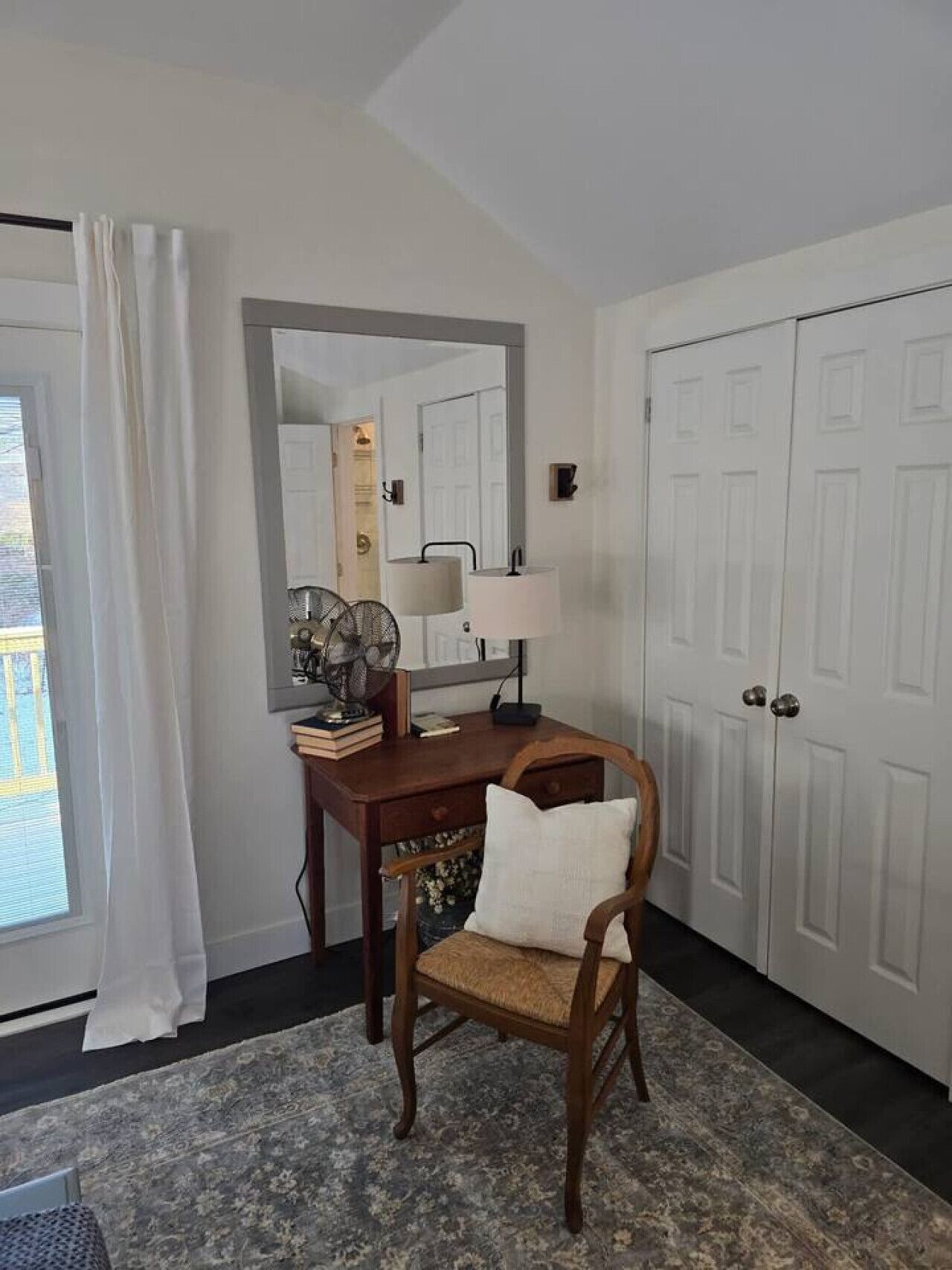 A room with a desk, chair, and a large mirror. There is a door with a light switch on the wall.