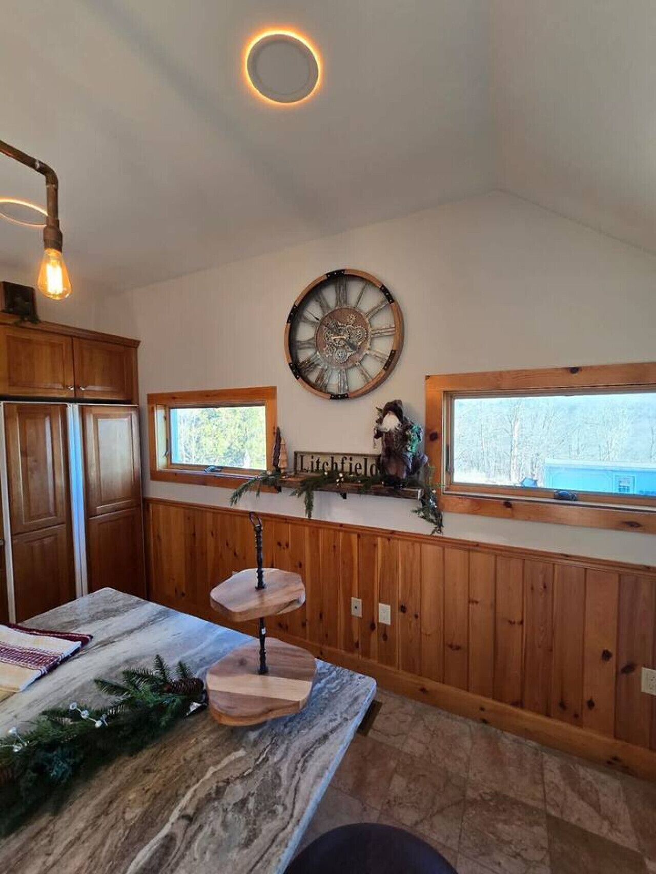 A kitchen with wooden cabinets, a marble countertop, a clock, and a window with a view of trees.
