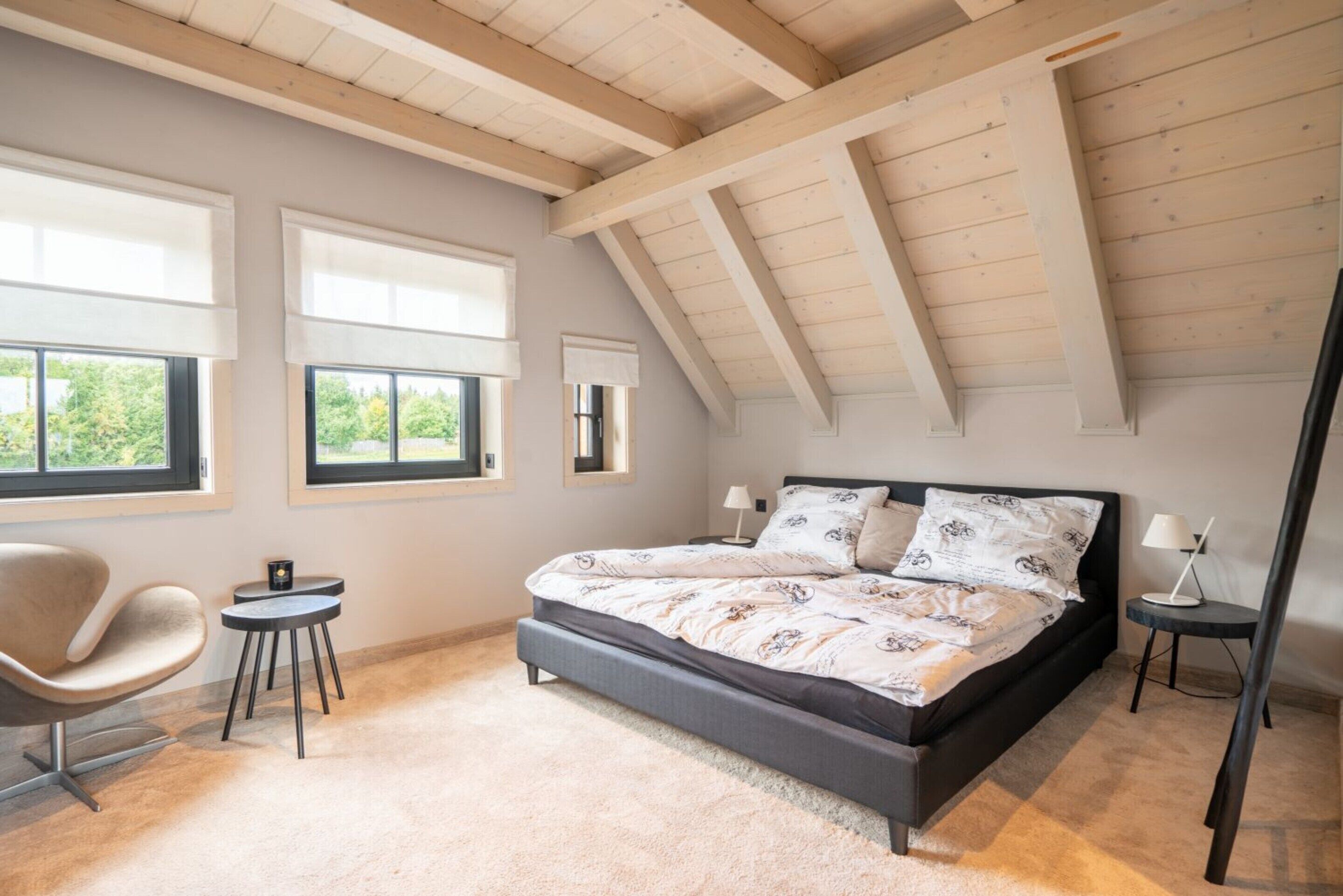 A bedroom with a sloped ceiling, a bed with patterned bedding, a chair, two side tables, and large windows with blinds.