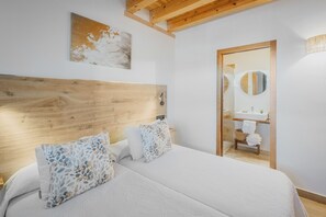 Un dormitorio con cabecera de madera, dos almohadas con estampado floral, una mesita de noche y vistas a un baño con espejo y lavamanos.