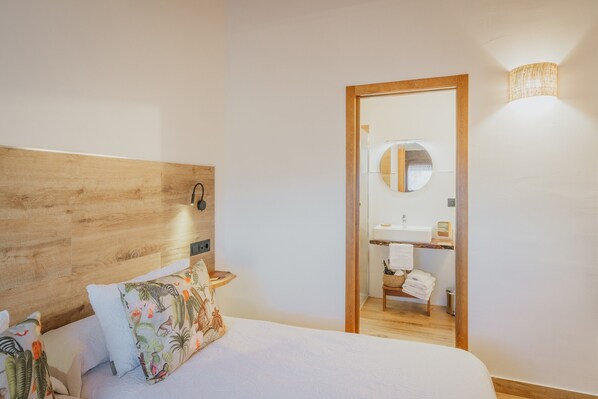 A bedroom with a wooden headboard, a bed with floral pillows, a bedside table, and a mirror reflecting a bathroom with a sink and towel rack.
