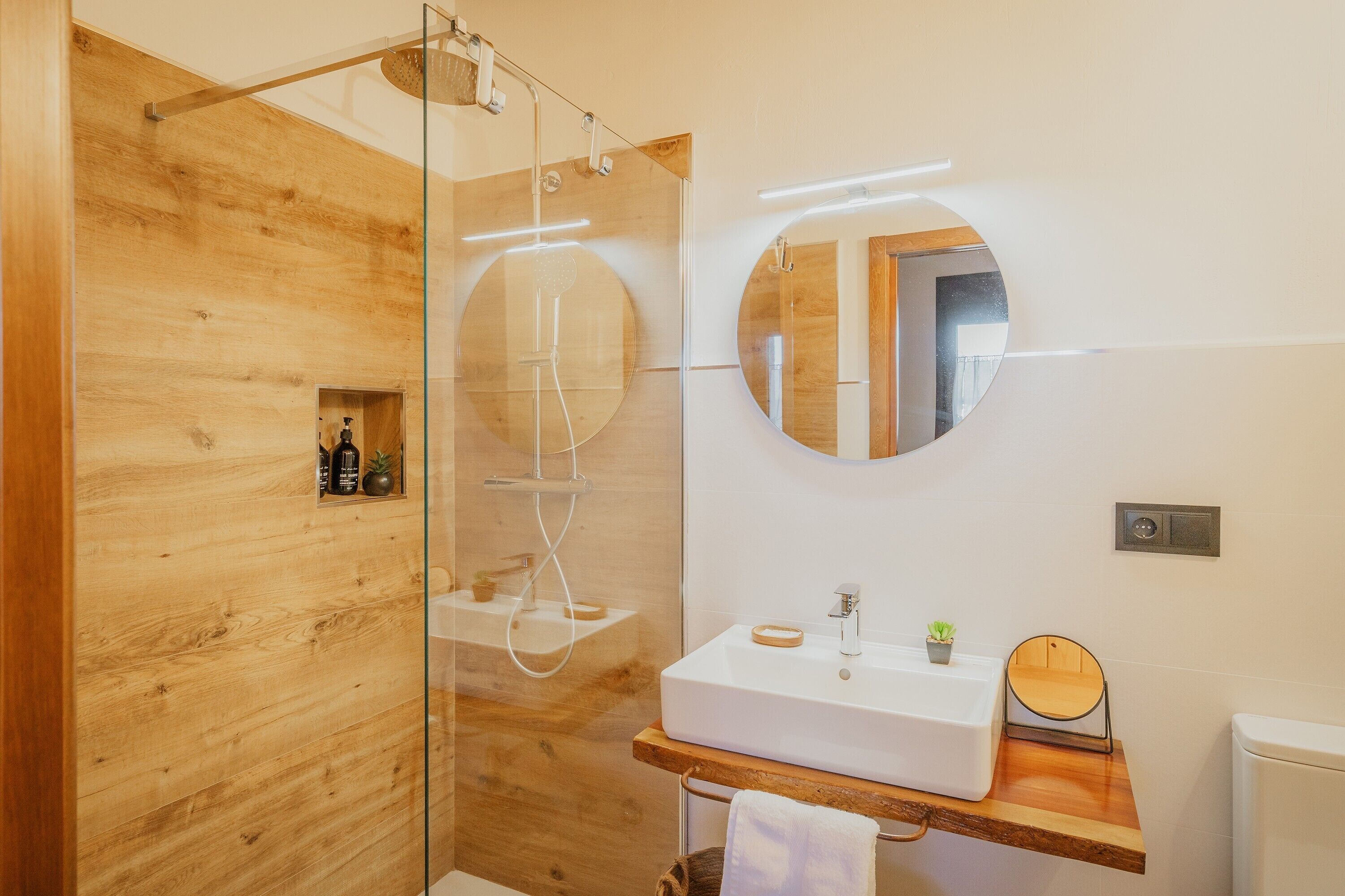 Een moderne badkamer met een houten wand, een douche met glazen wanden, een wit wastafelmeubel en een ronde spiegel.