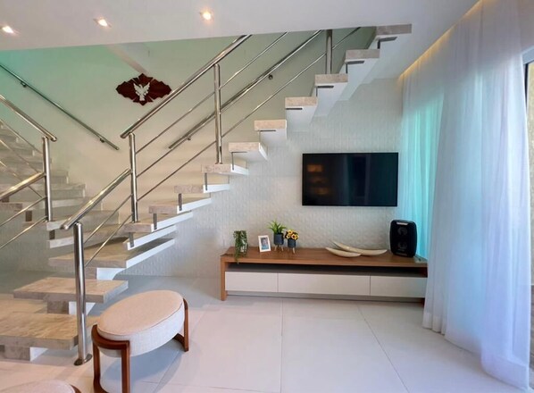 A modern living room with a staircase, a flat-screen TV, a wooden console table, and a bench.