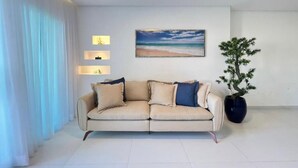 Een woonkamer met een beige bank, een plant in pot en een lijst met een zandstrand.