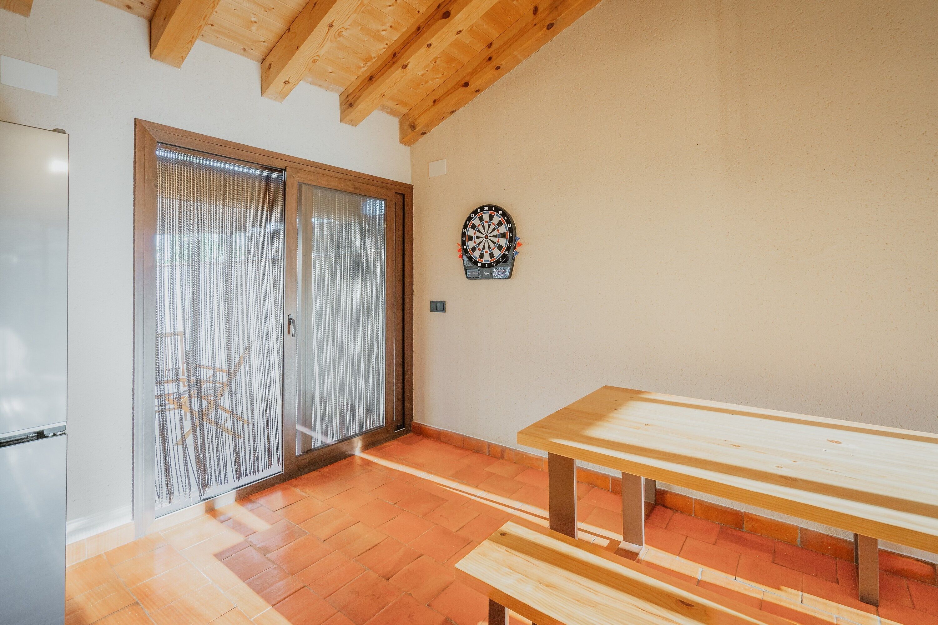 Une pièce avec un plafond en bois, un jeu de fléchettes, une porte-fenêtre coulissante et une table en bois.
