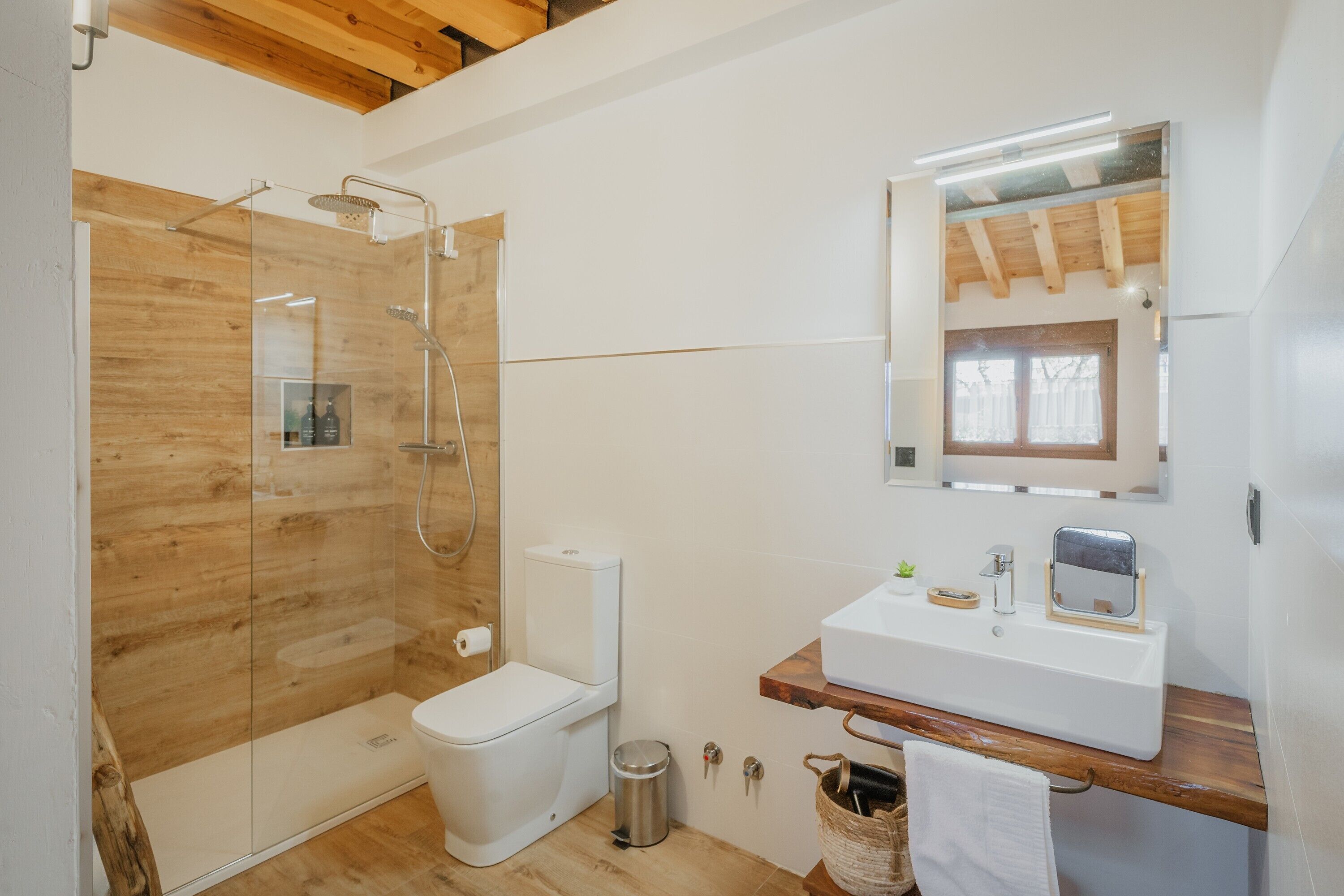 Un baño moderno con ducha de vidrio, inodoro blanco, tocador de madera con lavamanos y espejo.