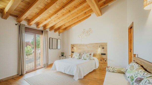 Ein Schlafzimmer mit Holzboden, einem Bett mit weißer Bettwäsche, einem Sofa mit Blumenmuster und Blick durch eine Glasdoor.