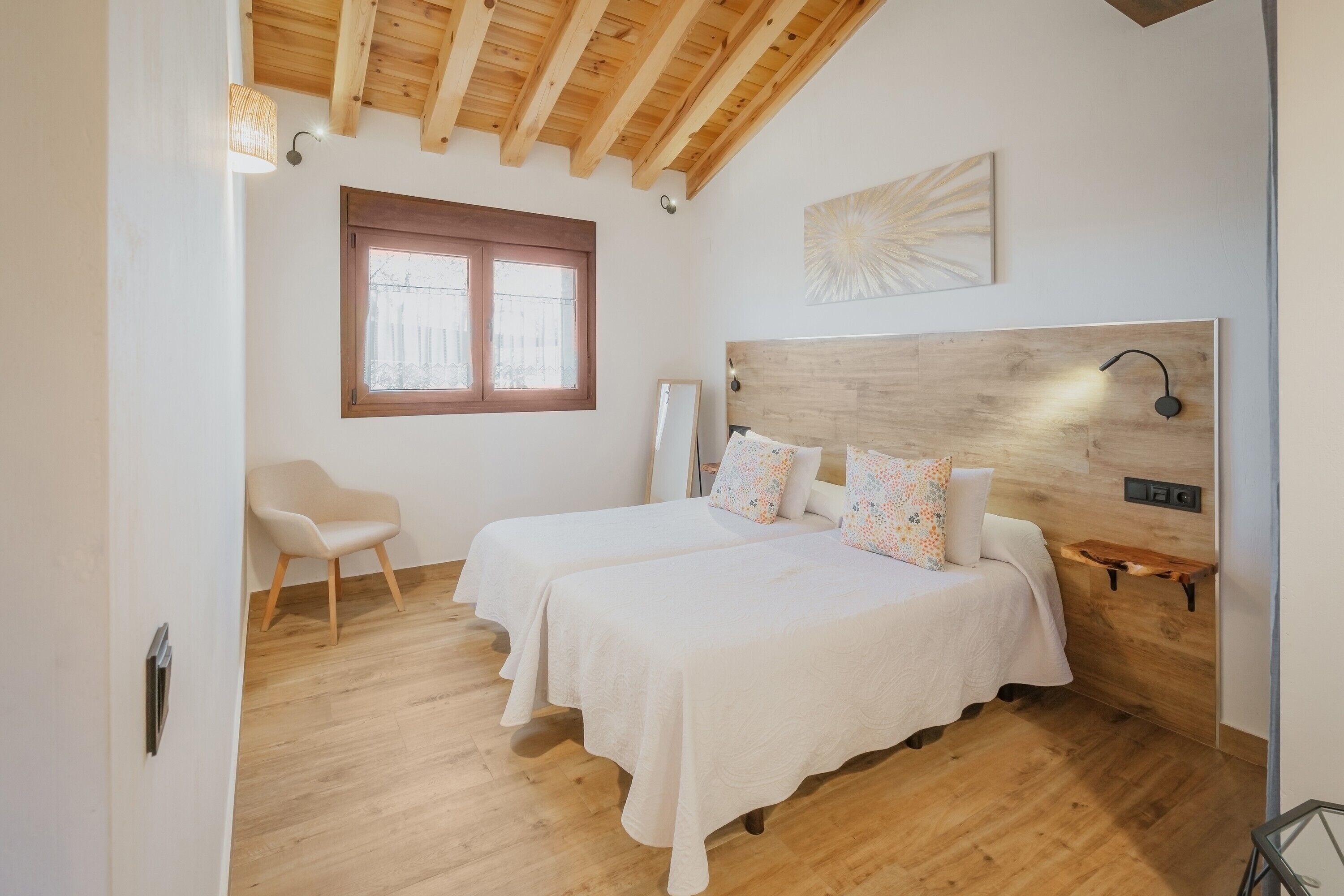 Un dormitorio con dos camas, techo de madera, una ventana, una silla y un estante en la pared.