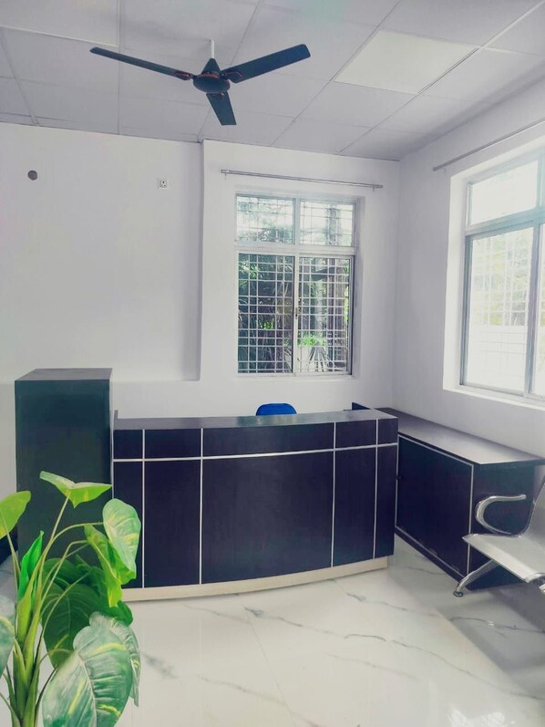 A reception area with a black reception desk, a ceiling fan, a window with metal bars, and a potted plant. - Hotel Silver Woods Ranchi (Ranchi)