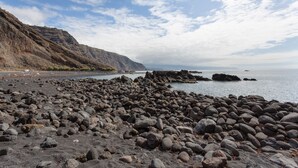 Una playa rocosa con grandes rocas, cielo despejado y montañas a lo lejos.