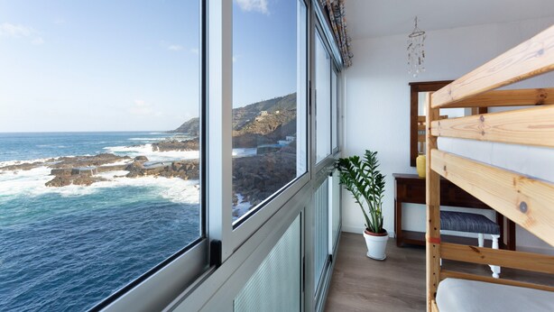 Habitación con un gran ventanal con vista a una costa rocosa, una cama litera de madera y una planta en maceta.
