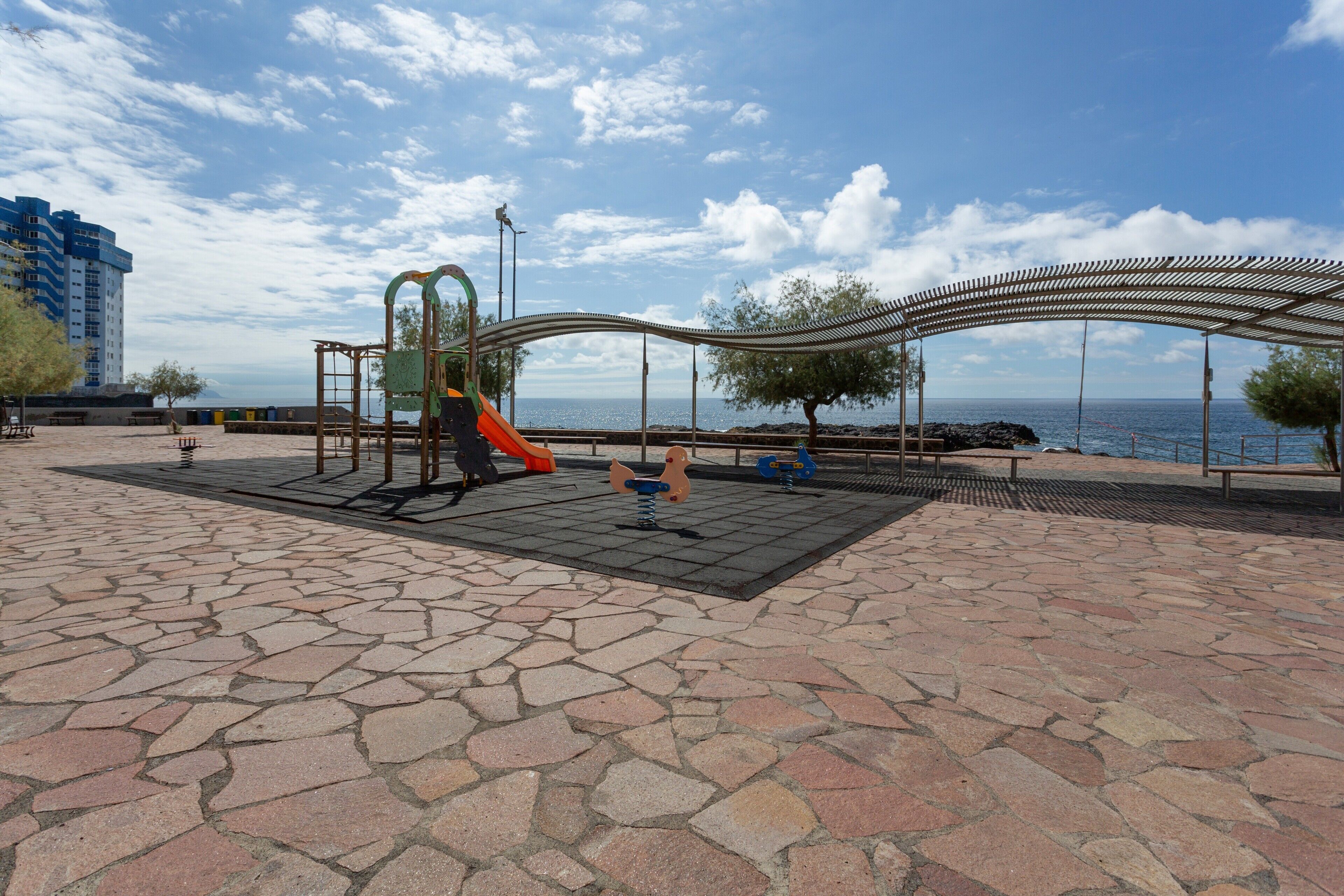 A playground with a slide and swings, a covered seating area, and a stone-paved ground.