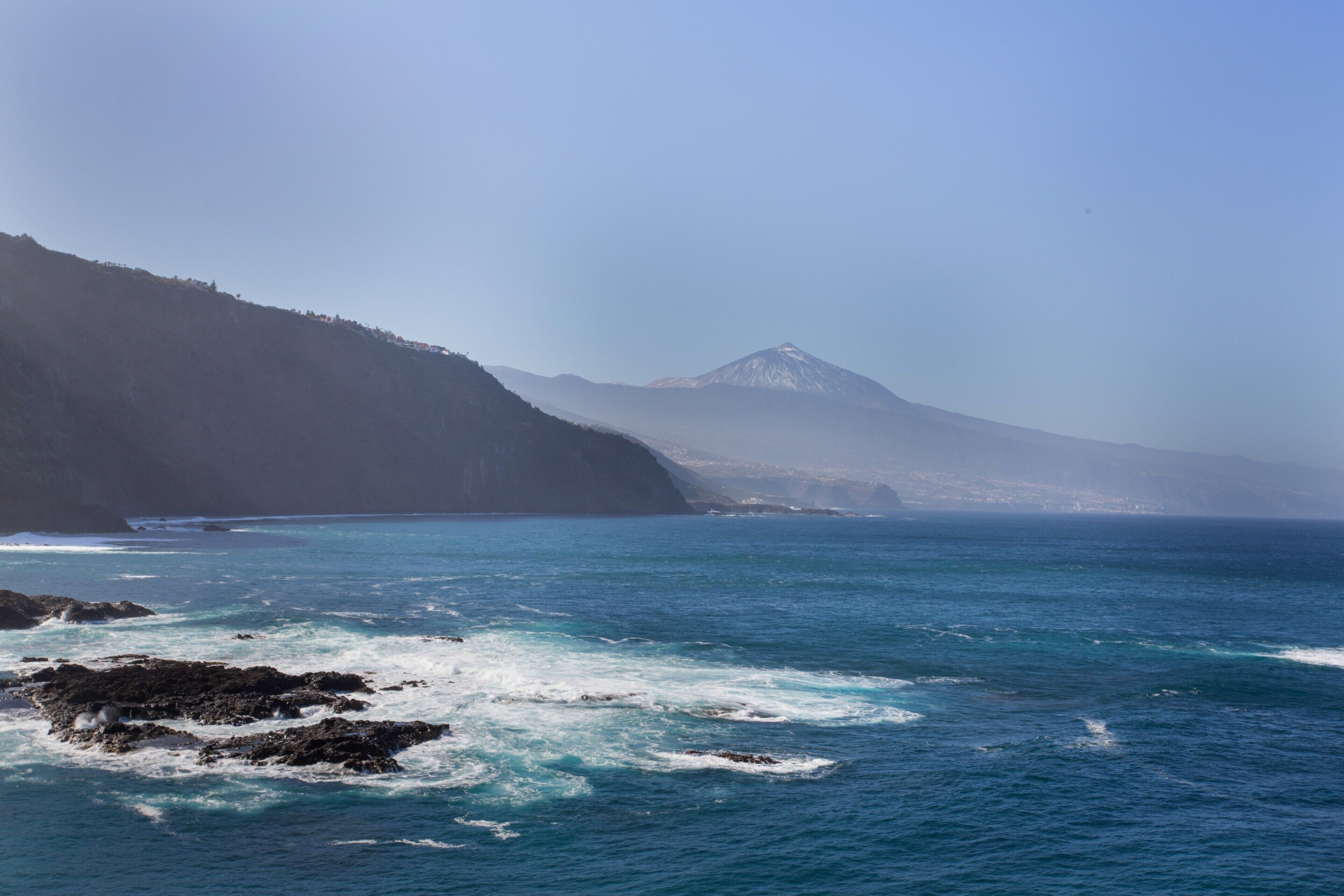 A coastal landscape with a mountain in the distance, rocky shoreline, and clear blue sea.