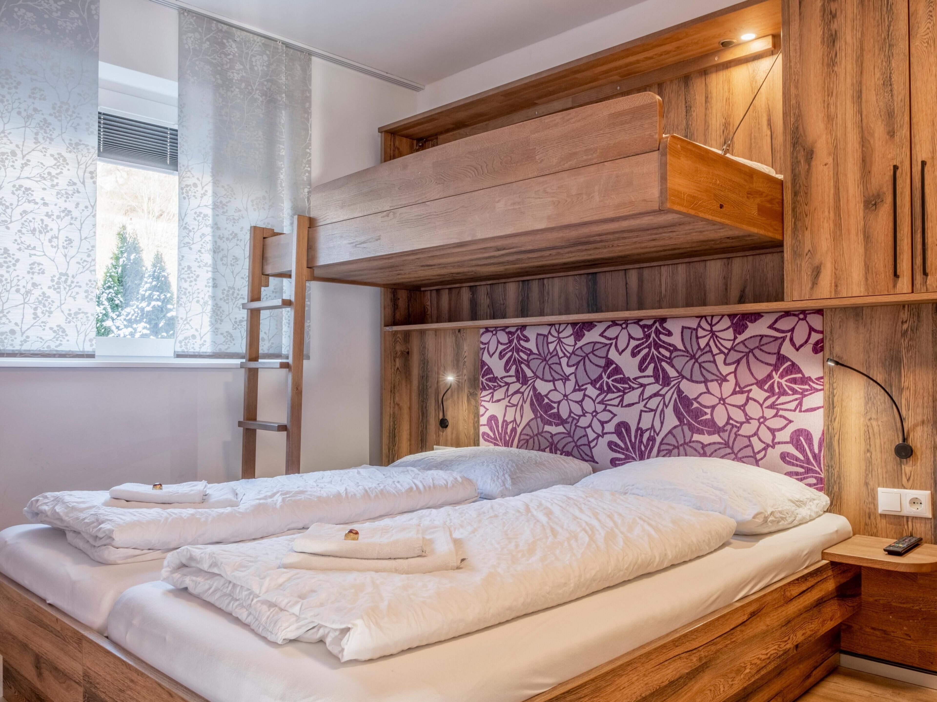 A bedroom with two beds, wooden headboard, a ladder, a window with a view of trees, and a wall-mounted light.