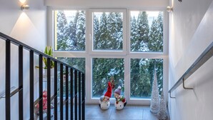 Un escalier avec une rampe, une vue sur des arbres couverts de neige à travers de grandes fenêtres et un petit sapin de Noël sur la plateforme.