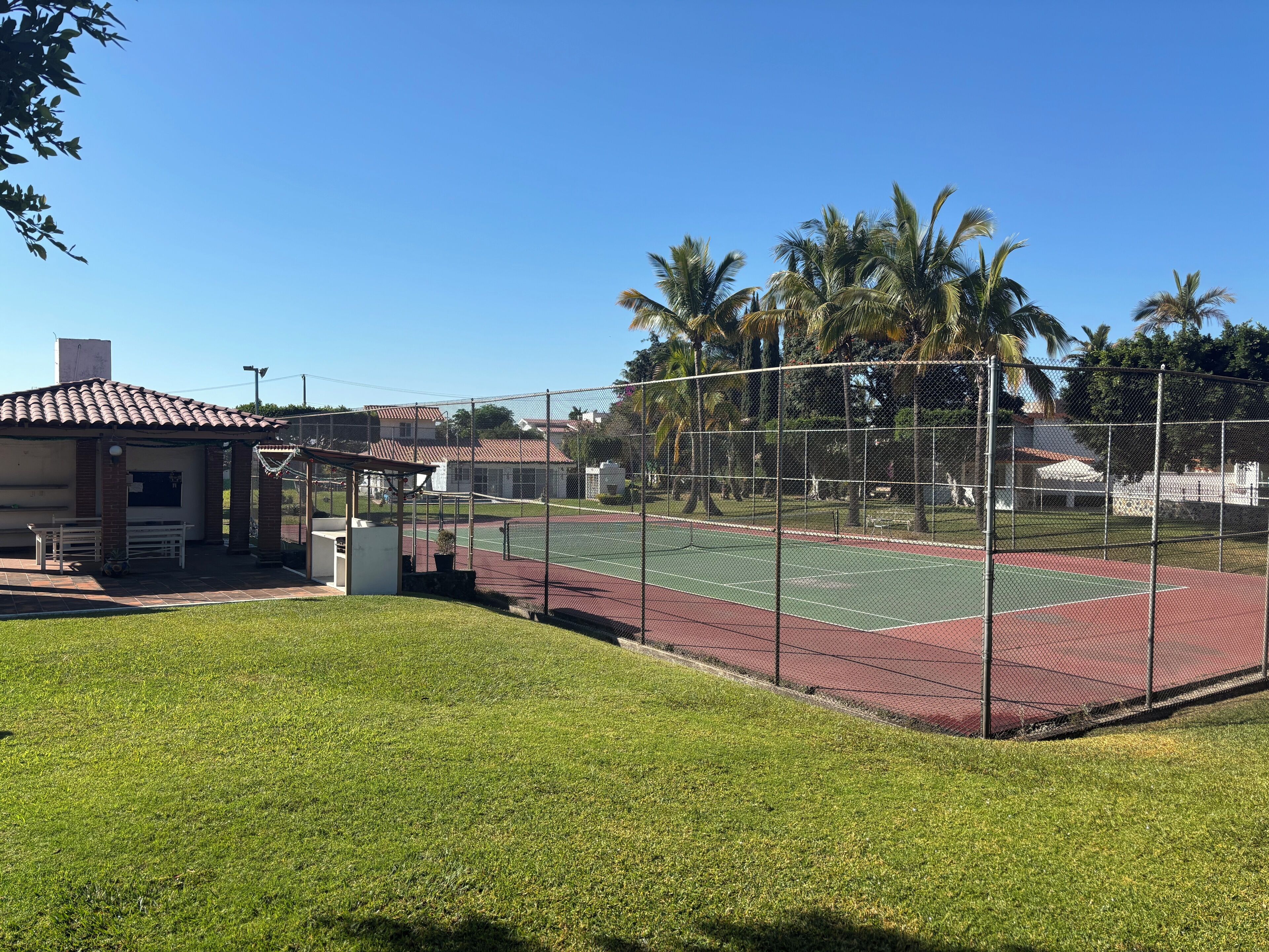 A tennis court with a fence, a small pavilion, and a grassy area.