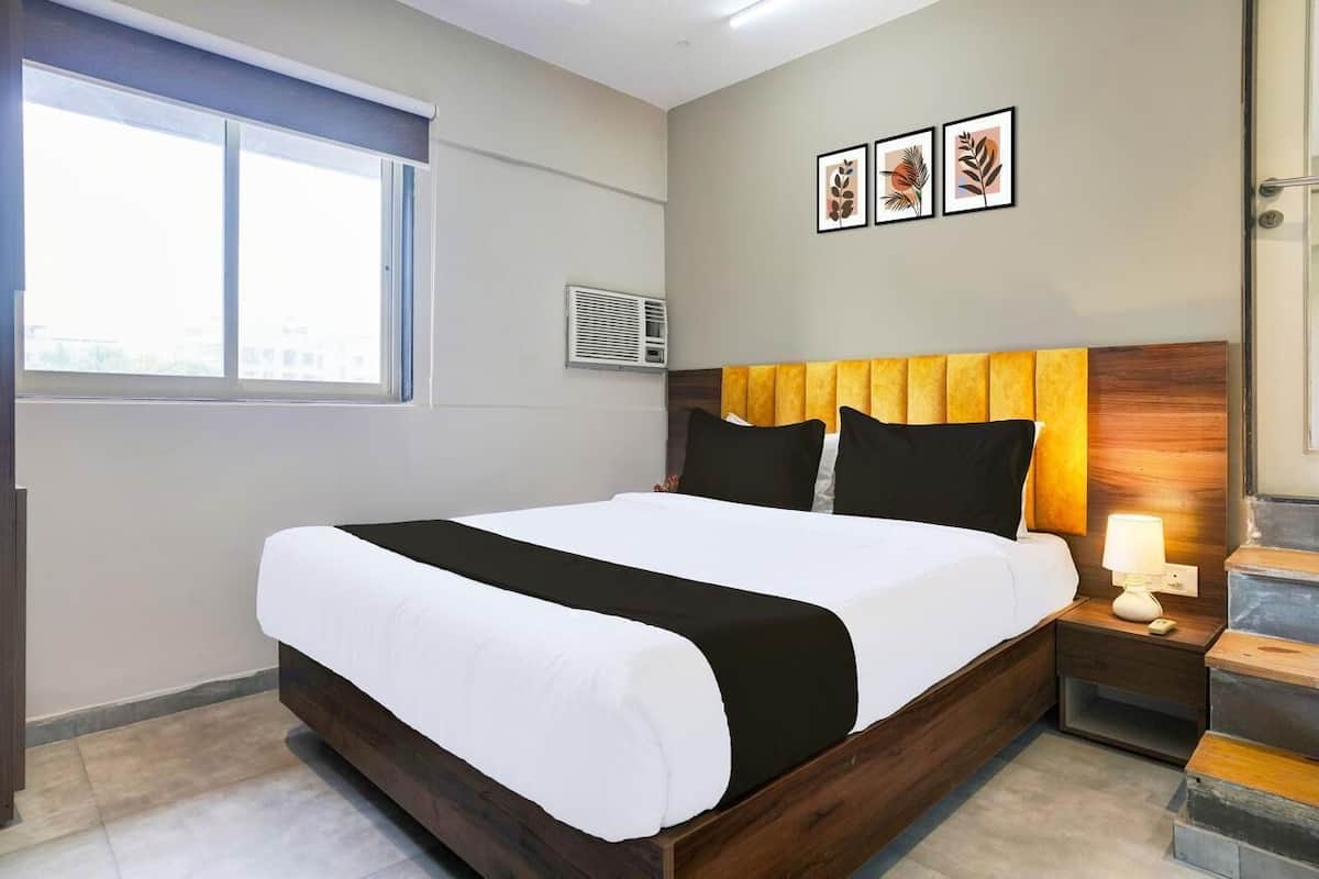 A hotel room with a bed, a window, an air conditioning unit, and a nightstand with a lamp.