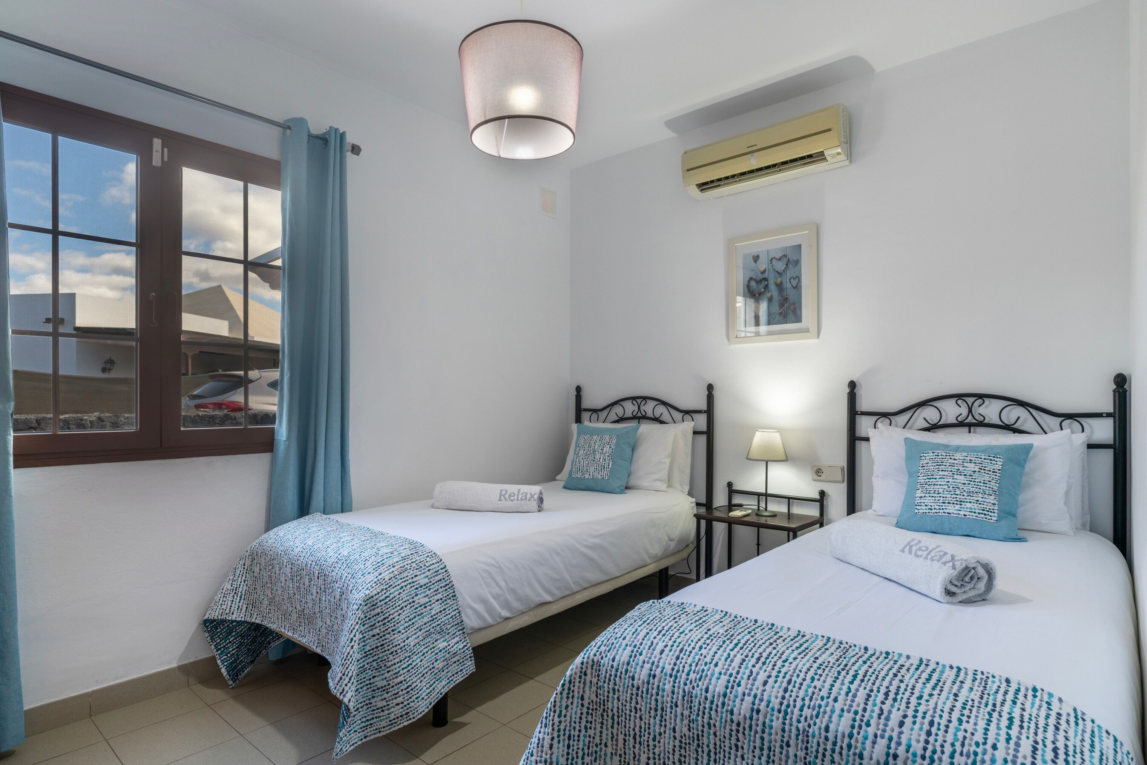 A bedroom with two beds, a window with curtains, a ceiling light, an air conditioner, and a framed picture on the wall.