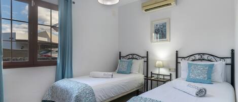 A bedroom with two beds, a window with curtains, a ceiling light, an air conditioner, and a framed picture on the wall.