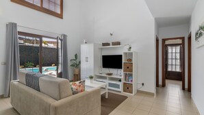 A living room with a sofa, TV, white cabinet, and a view of a swimming pool. - Vacation Villa 'Kalima' with Mountain View, Wi-Fi and Air Conditioning (Playa Blanca)