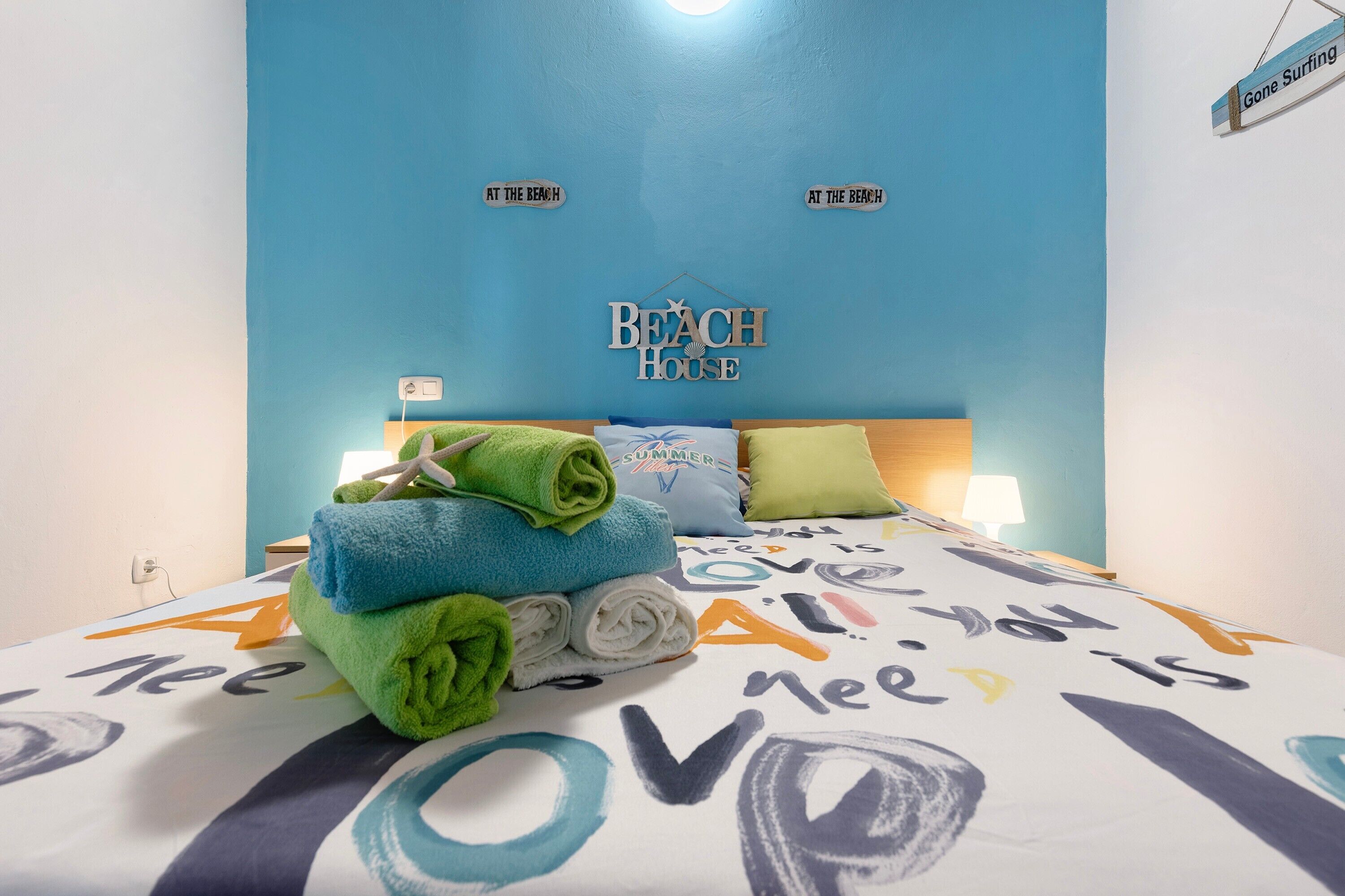 A bedroom with a blue wall, a bed with a colorful bedspread, and a sign that reads 'Beach House'.