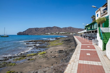 A coastal walkway with a restaurant, rocky beach, and mountains in the background.