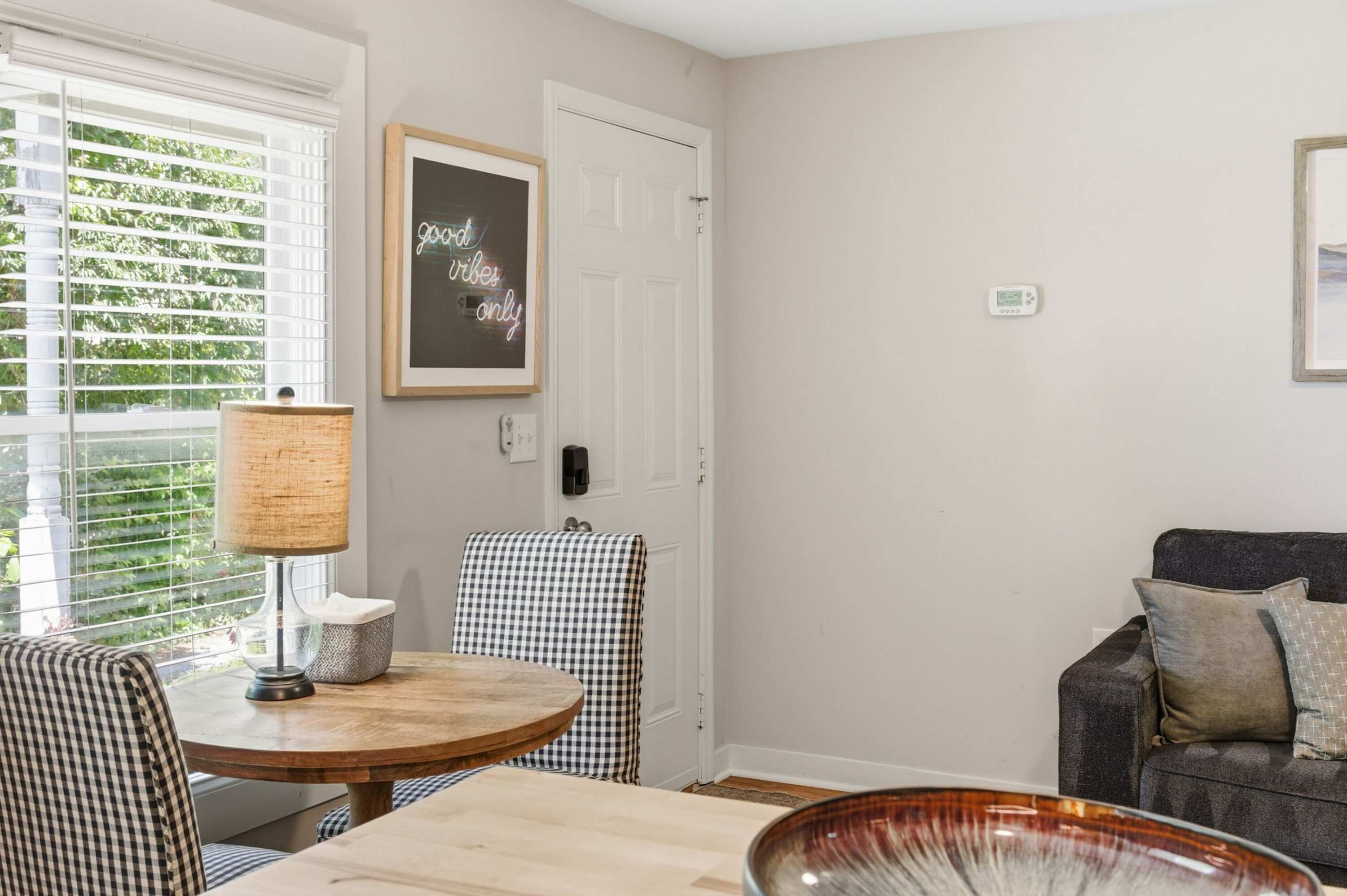 A room with a round wooden table, checkered chairs, a lamp, a framed sign, and a grey sofa.